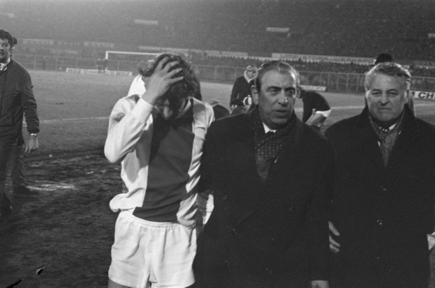 Eine Gruppe von Männern in Not steht zusammen auf einem Fußballfeld, mit einem Stadion im Hintergrund, in einem Schwarz-Weiß-Bild.