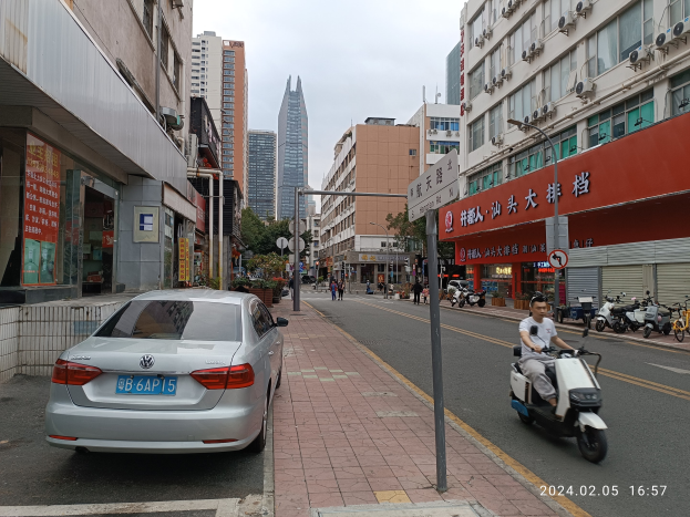 Ein Mann fährt mit einem Roller eine Straße mit hohen Gebäuden entlang, mit Menschen auf den Bürgersteigen, Fahrzeugen auf der Straße und Bäumen unter einem klaren blauen Himmel im Hintergrund.