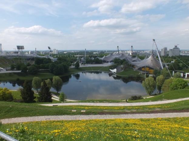 Ansicht des Olympiageländes von einem Hügel aus, mit einem Teich im Vordergrund, umgeben von Grünfläche und gelben Blumen, und Gebäuden im Hintergrund unter einem klaren blauen Himmel.