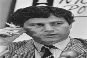 Ein Mann im Anzug und Krawatte raucht eine Zigarette, mit zwei Mikrofonen vor ihm und einer Tafel im Hintergrund, dargestellt in Schwarz-Weiß.