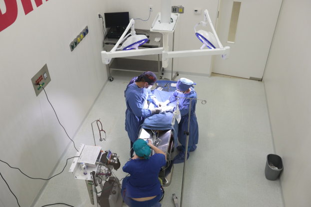 Eine Gruppe von Chirurgen führt einen Eingriff in einem Operationssaal durch, mit einem Patienten auf einer Trage umgeben von medizinischer Ausrüstung, Text an der linken Wand, einem Objekt auf dem rechten Boden und verschiedenen medizinischen Geräten im Hintergrund.