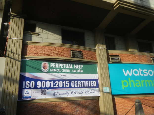 Außenansicht des Perpetual Help Medical Center - Las Pinas mit Säulen, Fenstern, Klimaanlagen, einem Banner mit seinem Namen und der ISO 9001:2015-Zertifizierung und einem Kettenzaun im Vordergrund.