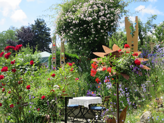 Ein farbenfroher Garten auf der Chelsea Flower Show mit bunten Blumen und Pflanzen, einem zentralen Tisch mit einer Decke und Stühlen, umgeben von Bäumen unter einem bewölkten Himmel.