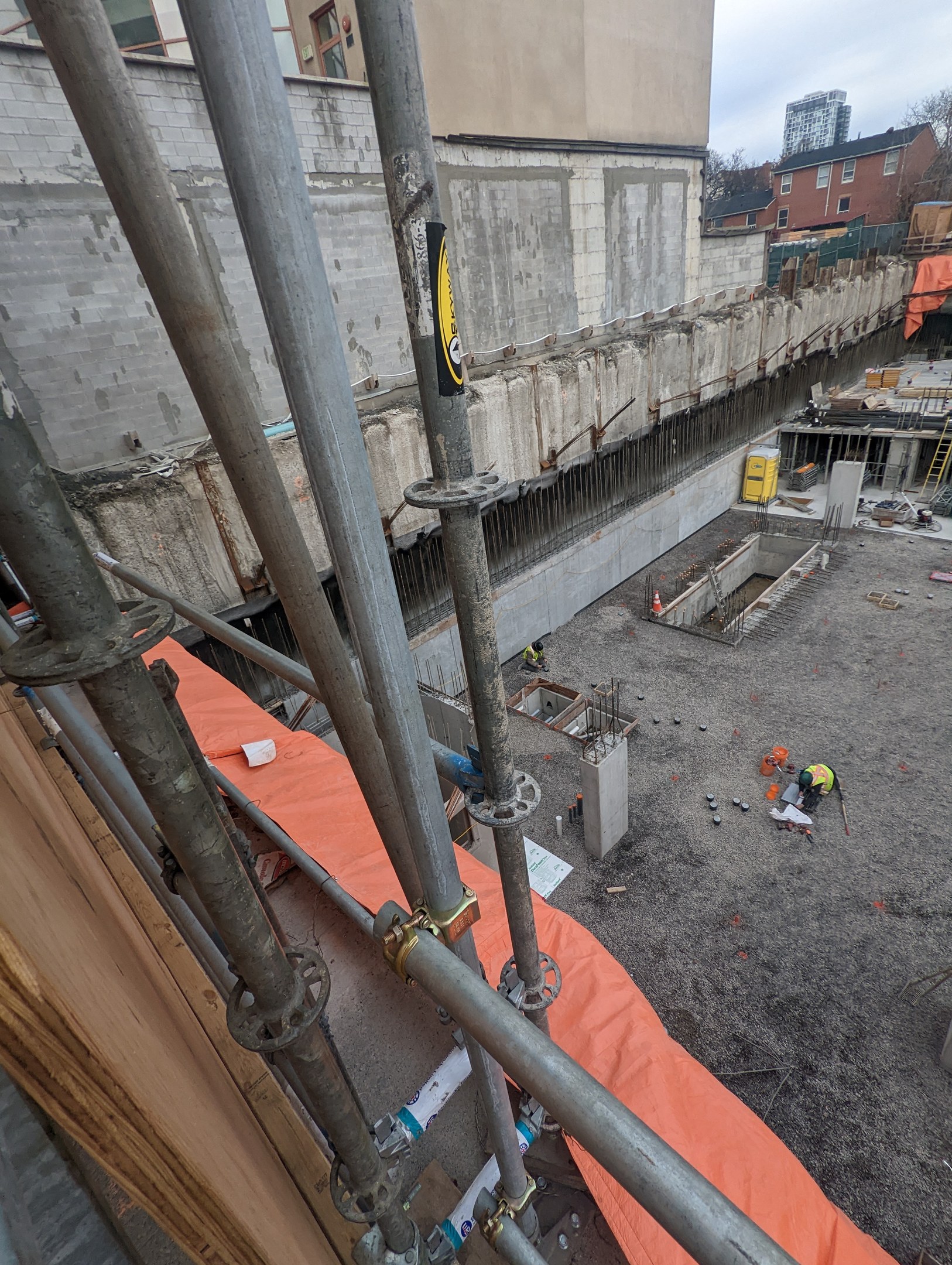 Baustelle mit einem großen Gebäude in Renovierung im Hintergrund, umgeben von Bäumen und einem klaren blauen Himmel, und Arbeitern, die Metallstangen und Rohre im Vordergrund bearbeiten.