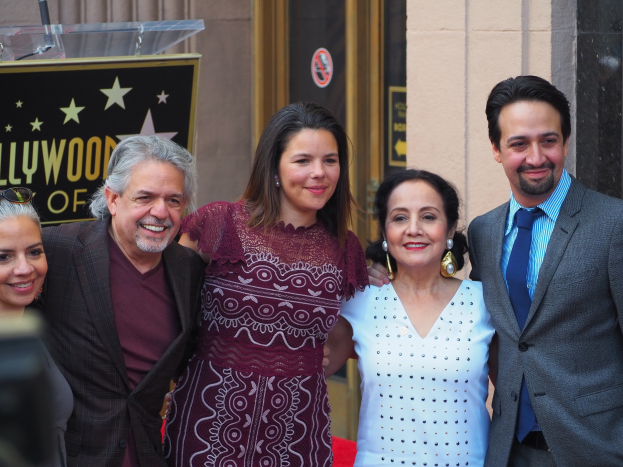 Eine Gruppe von Menschen posiert auf einem roten Teppich bei der Zeremonie des Hollywood Walk of Fame, mit einer Kamera auf der linken Seite, einer Tafel mit Text, einem Glaspodest und einer Wand im Hintergrund.