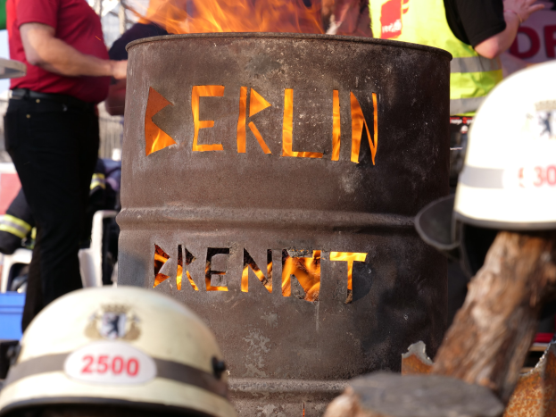 Eine Gruppe von Menschen steht in der Nähe eines Feuerhydranten mit der Aufschrift "Berlin", mit einem brennenden Fässer im Vordergrund, Helme unten und ein hölzernes Objekt rechts.