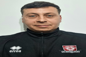 Ein Mann in einer schwarzen Jacke und einem T-Shirt mit einem Logo des Croydon Athletic Football Club steht vor einer weißen Wand.