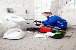 Ein Mann in einer blauen Uniform sitzt auf dem Boden vor einem Urinal und hält ein Rohr in der Hand, umgeben von Reinigungswerkzeugen und -mitteln, mit Urinalen auf der linken und einem Fenster auf der rechten Seite.