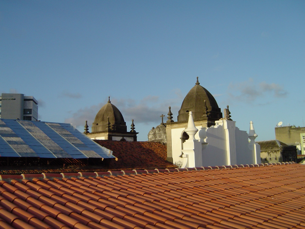 Stadtansicht mit Gebäuden im Vordergrund und einem blauen Himmel im Hintergrund, mit Solarpanelen auf einem Dach, die den Einsatz erneuerbarer Energie anzeigen.