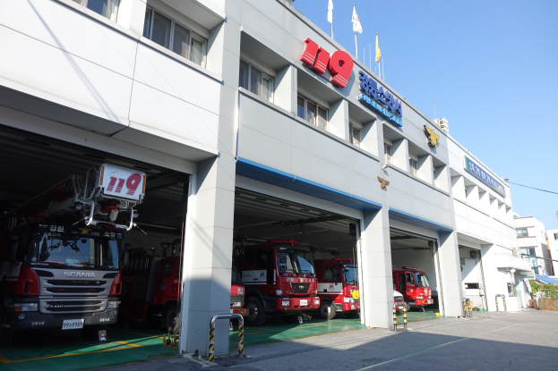 Feuerwache mit mehreren Feuerwehrautos vor dem Gebäude mit Fenstern, Säulen und Namensschildern, mit Fahnen im Vordergrund und anderen Gebäuden gegen einen klaren blauen Himmel.