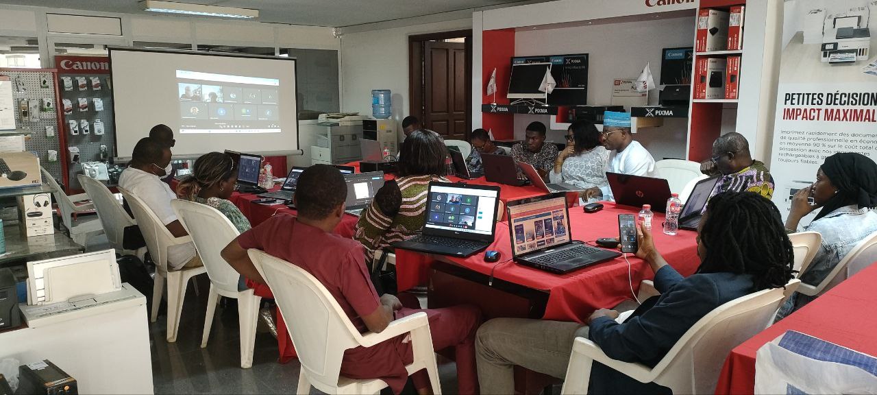 Gruppe von Menschen, die um einen Tisch mit Laptops und anderen Gegenständen während eines digitalen Marketing-Workshops sitzen, mit einem Banner, einem Display, einer Tür und Deckenleuchten im Hintergrund.