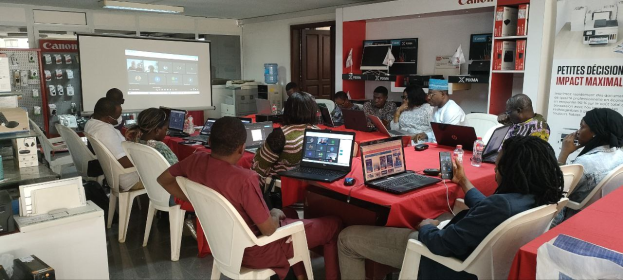 Gruppe von Menschen, die um einen Tisch mit Laptops und anderen Gegenständen während eines digitalen Marketing-Workshops sitzen, mit einem Banner, einem Display, einer Tür und Deckenleuchten im Hintergrund.