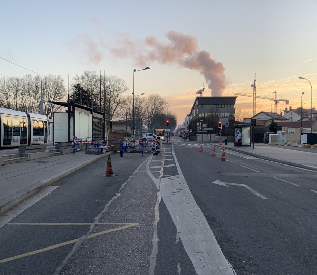 Eine Stadtstraße mit Verkehrskegeln, einem Bus, Laternenmasten, Bäumen, Gebäuden, Kränen und Rauch, der aus einem fernen Gebäude aufsteigt, vor einem Himmel.