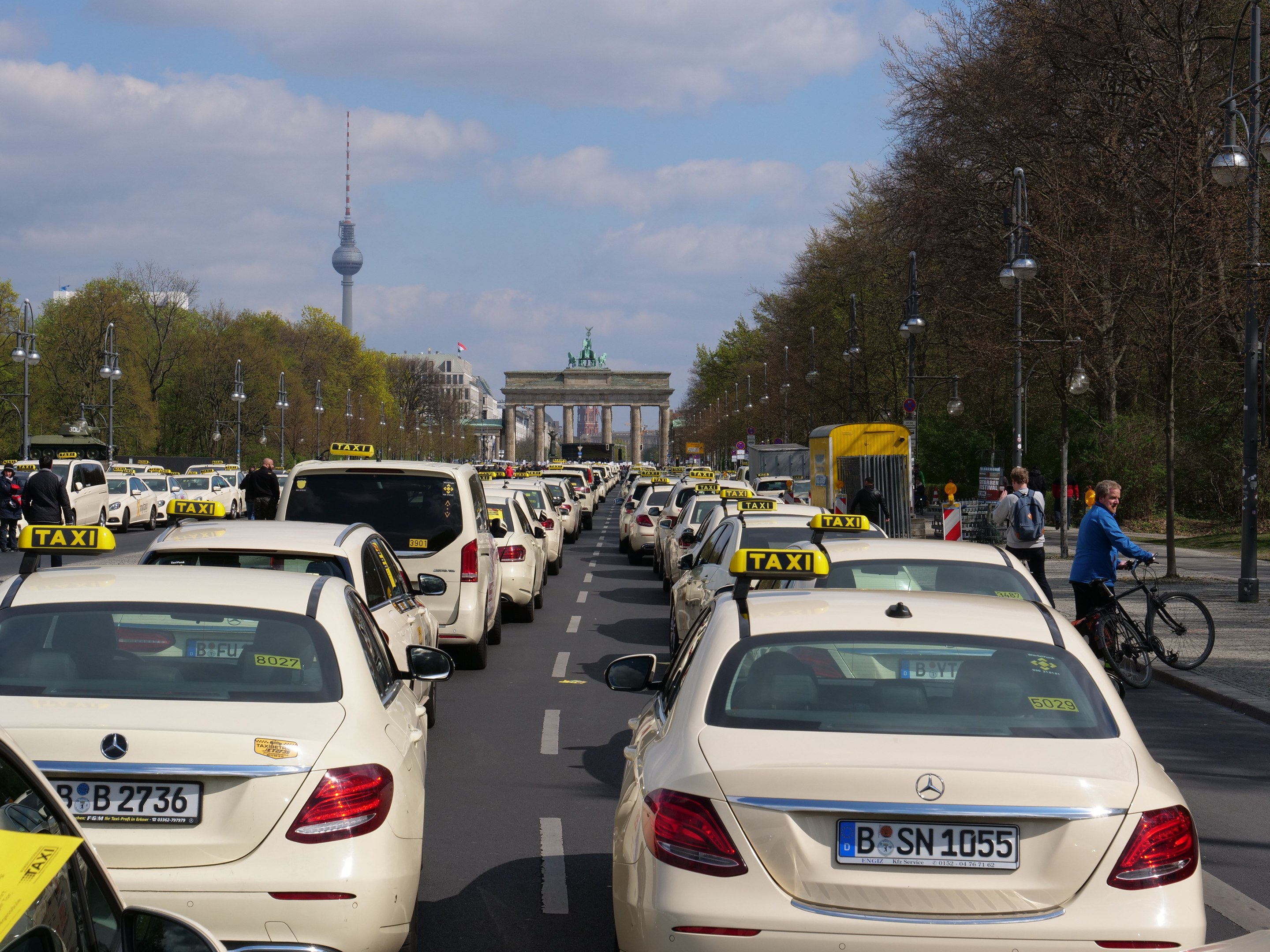Eine lange Reihe von Taxis, die entlang einer belebten Straße in Berlin, Deutschland, geparkt sind, mit Fahrzeugen, Fahrradfahrern und Fußgängern in der Nähe, flankiert von Laternenmasten, Bäumen und Gebäuden, mit einem Bogen und einem Turm im Hintergrund unter einem bewölkten Himmel.