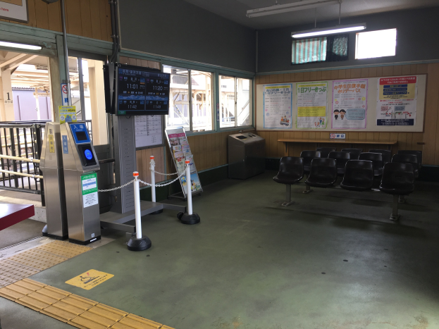 Ein belebter Bahnhofseingang mit Sitzgelegenheiten, einem Service-Schalter, Ticketautomaten, Kettenbarrieren, Informationsanzeigen, Wandplakaten, Deckenleuchten und Geländern.