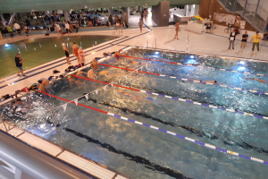 Ein großes Hallenbad mit schwimmenden, stehenden und sitzenden Menschen, Treppen und Geländern auf der rechten Seite und einer Wand im Hintergrund.