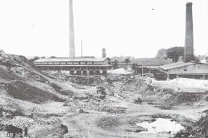 Ein Schwarz-Weiß-Foto einer Baustelle mit zwei Schornsteinen im Hintergrund, umgeben von Gebäuden, Masten, Bäumen und einem klaren Himmel, mit Text am unteren Rand, der darauf hinweist, dass es sich um ein Kohlekraftwerk handelt.