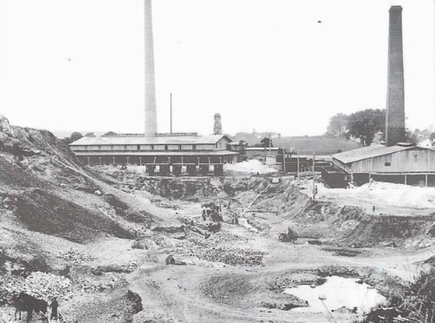 Ein Schwarz-Weiß-Foto einer Baustelle mit zwei Schornsteinen im Hintergrund, umgeben von Gebäuden, Masten, Bäumen und einem klaren Himmel, mit Text am unteren Rand, der darauf hinweist, dass es sich um ein Kohlekraftwerk handelt.