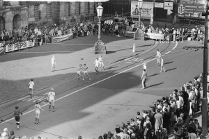 Schwarzes Foto eines auf der Straße ausgetragenen Fußballspiels mit Zuschauern, einige stehen, einige sitzen, mit Laternenmasten, Gebäuden und Transparenten an der Straße.