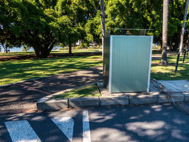 Transportable Toilette an der Seite einer Straße in einem Park, umgeben von Bäumen und Gras, mit einem Gewässer und einem klaren blauen Himmel im Hintergrund.
