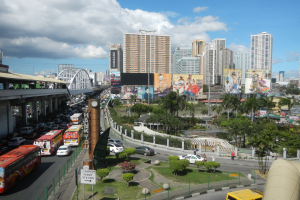 Eine belebte Stadtstraße mit Bussen, Autos und anderen Fahrzeugen, umgeben von Bäumen, Pflanzen, Gras, Pfählen, Plakaten, einer Brücke, Gebäuden und einem bewölkten Himmel.