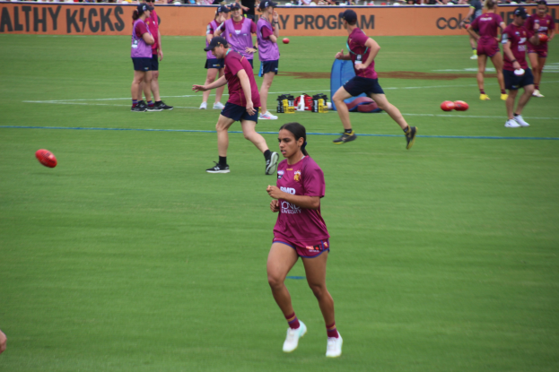 Frauen spielen Australian Rules Football auf einem grünen Feld mit verstreuten Bällen, einige tragen Kappen und Turnschuhe und ein Banner mit Text im Hintergrund.