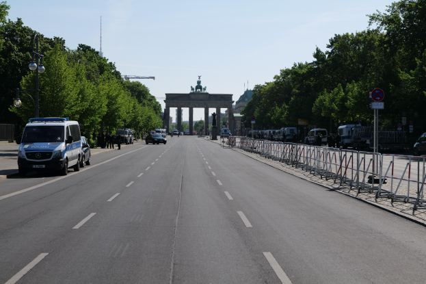 Ein Polizeiwagen steht auf der Seite einer vielbefahrenen Straße vor dem Brandenburger Tor in Berlin, Deutschland, mit Barrieren, Schildern, Bäumen und Laternenmasten im Hintergrund und einer bewölkten Himmel.