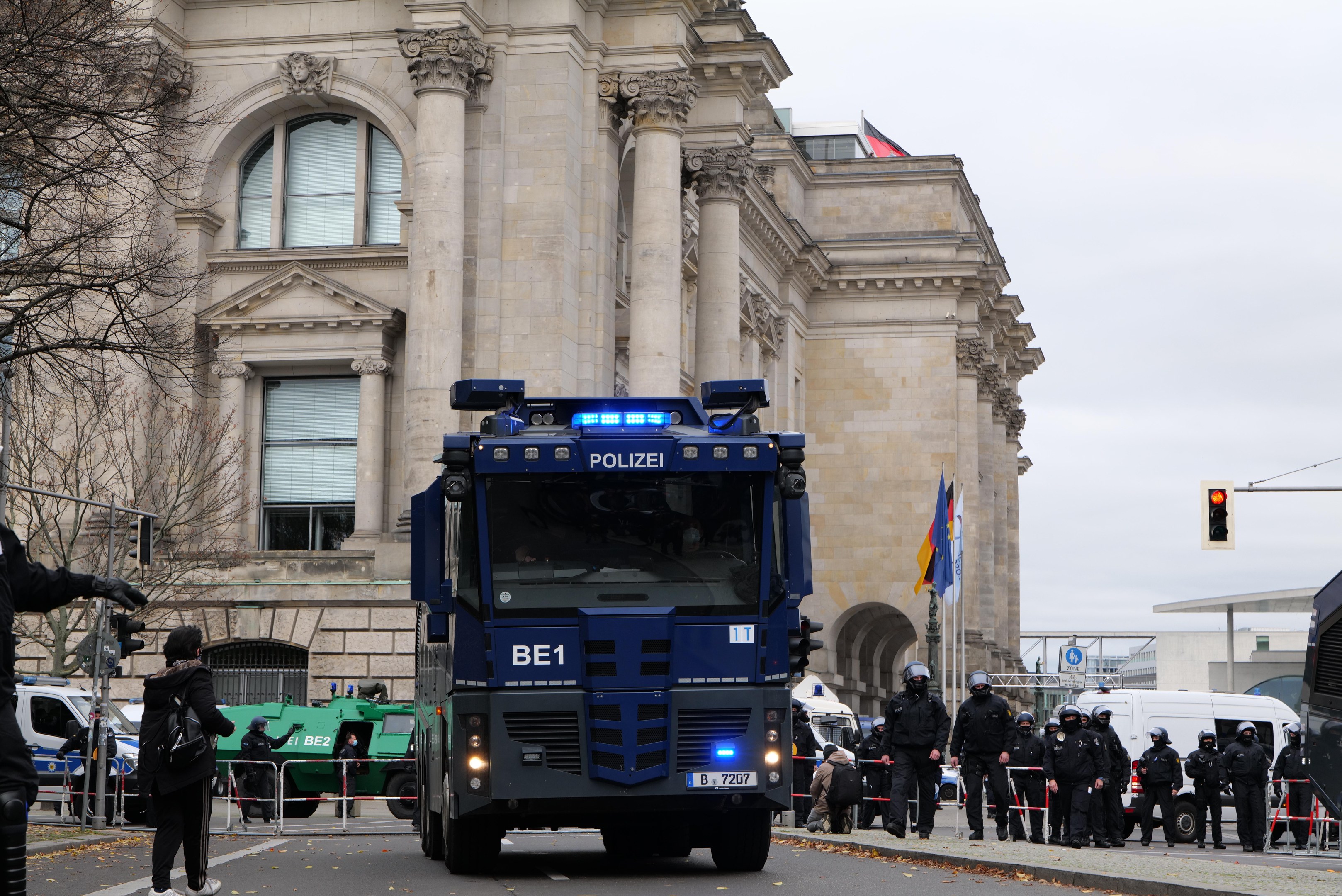 Gruppe von Polizisten vor einem großen Gebäude mit architektonischen Details, Fahrzeuge auf der Straße, eine Person mit einer Kamera links, Bäume, Verkehrsampeln, Fahnenmasten und einen klaren blauen Himmel.