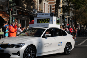 Ein weißer BMW mit einer Uhr auf dem Dach fährt eine Straße entlang, mit einer Person im Auto und einer Gruppe von Menschen am Straßenrand, während Gebäude, Bäume und Verkehrszeichen im Hintergrund zu sehen sind.
