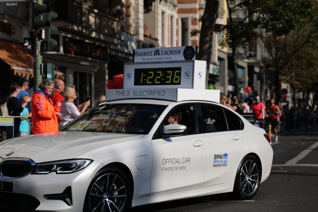Ein weißer BMW mit einer Uhr auf dem Dach fährt eine Straße entlang, mit einer Person im Auto und einer Gruppe von Menschen am Straßenrand, während Gebäude, Bäume und Verkehrszeichen im Hintergrund zu sehen sind.