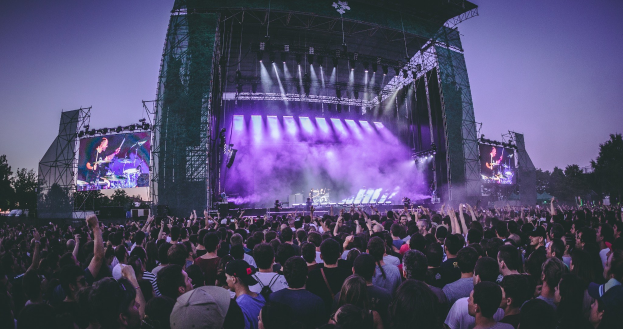 Eine große Menschenmenge auf einem Musikfestival mit einer Bühne, auf der Musiker Instrumente spielen, beleuchtet von Lichtern und umgeben von Bildschirmen, vor einer Kulisse aus Gebäude, Bäumen und einem klaren blauen Himmel.