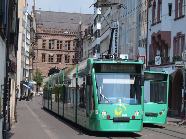 Zwei grüne Straßenbahnen fahren eine von hohen Gebäuden gesäumte Stadtstraße entlang, mit parkenden Fahrrädern und Fußgängern auf den Gehwegen, unter einem klaren blauen Himmel mit einem Baum im Hintergrund.