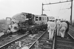 Ein Schwarz-Weiß-Bild einer Zugkollision mit einer Gruppe von Menschen in Helmen drumherum, mit Strommasten, Drähten, Bäumen, Gebäuden und einem klaren Himmel im Hintergrund.
