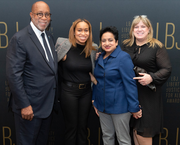 Vier Personen auf einem roten Teppich, die lächeln und für ein Foto posieren, mit einem 'Liberty Justice for All Awards'-Schild im Hintergrund.