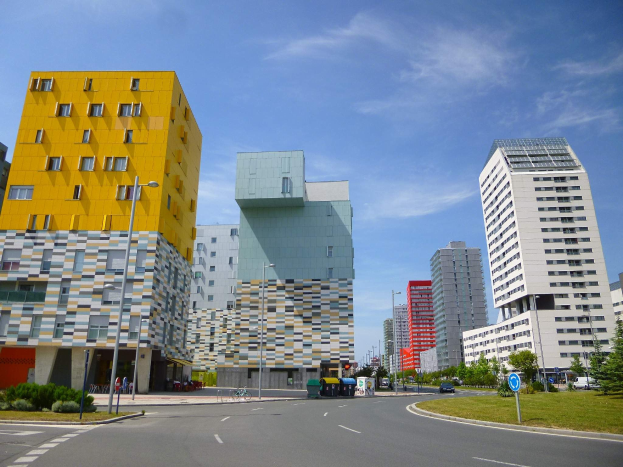 Eine Stadtstraße mit hohen Gebäuden im Hintergrund, Straßenlaternen, Straßenlaternen, Kraftfahrzeugen, Informationsschildern, Büschen, Bäumen und einem bewölkten Himmel, mit den einzigartig gestalteten Gebäuden der Universität Rotterdam.