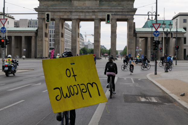 Eine Gruppe von Radfahrern fährt an der Brandenburgertor in Berlin vorbei, einer hält ein gelbes Schild, mit Laternenpfählen, Verkehrszeichen, Gebäuden, Bäumen und einem klaren Himmel im Hintergrund.