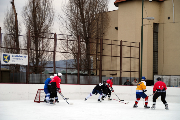 Menschen, die Eishockey auf einem Eisplatz mit Gebäuden, Bäumen, einer Straßenlaterne, einem Namensschild und Zäunen im Hintergrund unter einem Himmel spielen.