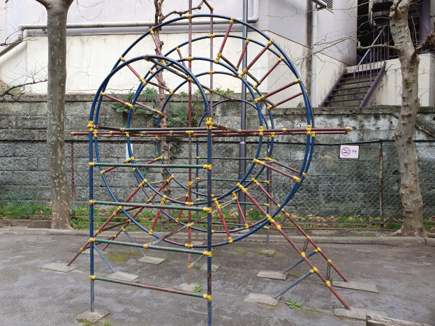 Ein Spielplatz mit einem großen Metallklettergerüst, umgeben von Bäumen und einer Maschendrahtzaun, mit Treppen mit Geländern, die zu einem Gebäude mit Fenstern und Rohren führen, einem Schild an einem Pfahl und grasbewachsenem Boden.