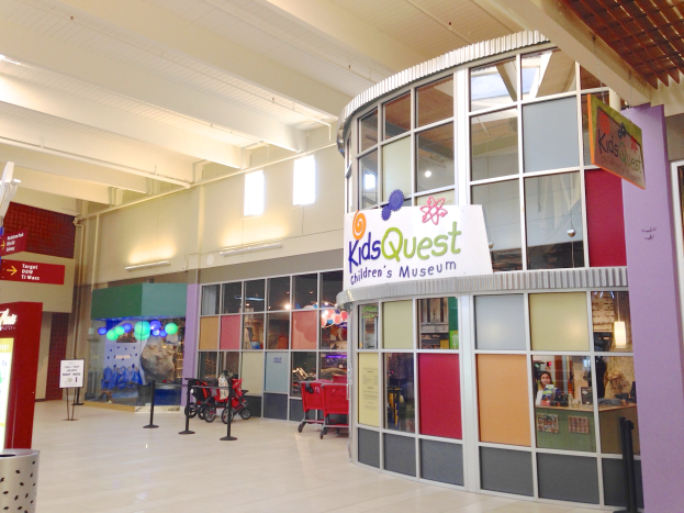 Innenansicht eines Einkaufszentrums mit einem "Kids Quest Children's Museum"-Schild, mit Einkaufswagen, ribboned poles, text boards, ceiling lights und verschiedenen verstreuten Gegenständen.