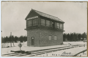 Ein Schwarz-Weiß-Foto einer Signalbox auf Eisenbahnschienen, umgeben von Bäumen und Masten, mit einer sichtbaren Tür, Fenstern und dem Himmel im Hintergrund.
