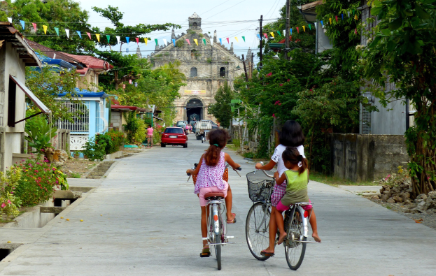 Drei Kinder fahren Fahrräder eine von Bäumen gesäumte Straße entlang mit Gebäuden auf beiden Seiten, einem Auto und einem Gebäude im Hintergrund und dem Himmel darüber.