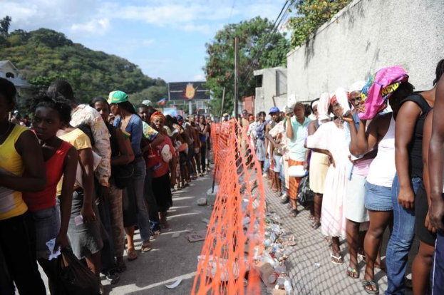 Lange Schlange von Menschen, die an einer Wahllokalwarteschlange stehen, mit einem Zaun, einer Wand, Bäumen, Gebäuden, Strommasten, Drähten, einem Plakat und Himmel im Hintergrund.