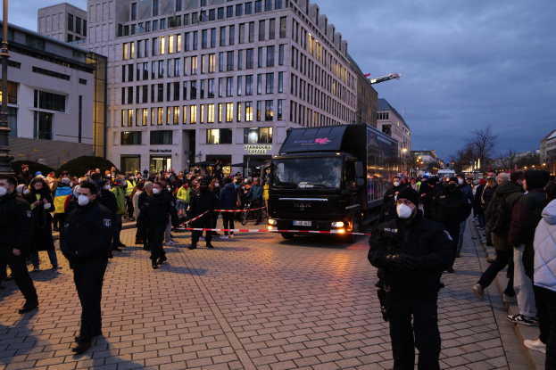Eine Gruppe von Menschen steht in der Nähe eines Lastwagens auf einer Straße mit Gebäuden, Bäumen und Laternen im Hintergrund, einige tragen Mützen und Masken, mit einem Band an einem Pfahl im Vordergrund.