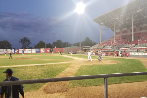 Baseballspiel in einem Stadion mit Zuschauern auf den RÀngen, GelÀnder im Vordergrund, BÀume, MÀste, Lichter, Werbetafeln und wolkenlosem Himmel im Hintergrund.