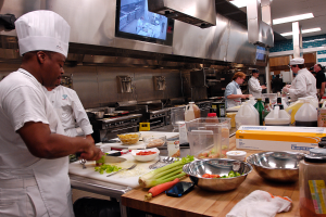 Eine Gruppe von Köchen in einer Küche, die auf einem Schneidbrett Lebensmittel vorbereiten, wobei ein Koch im Vordergrund eine weiße Schürze und eine Haube trägt, ein Messer hält und von verschiedenen Utensilien, Schüsseln, Gemüse, einem Mobiltelefon, Flaschen und anderen Gegenständen umgeben ist, während im Hintergrund ein Fernseher zu sehen ist.