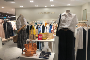 Ein Vanessa Bruno-Laden in Singapur mit Schaufensterpuppen in Kleidern, hängenden Kleidern, Taschen, einem Textschild an der Wand, einem Spiegel auf der rechten Seite und Deckenleuchten.