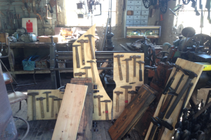 Eine Werkstatt voller Werkzeuge, Ausrüstung und Holzplanken, mit einem Stuhl links, Werkzeugen an der Wand und einem Fenster im Hintergrund.