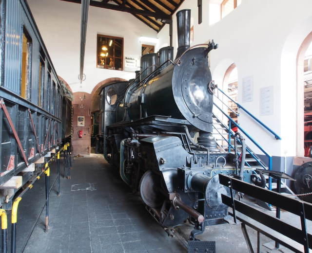 Alte Dampflokomotive drinnen in einem Museum präsentiert, umgeben von Informationsschildern, Geländern und Beleuchtung.