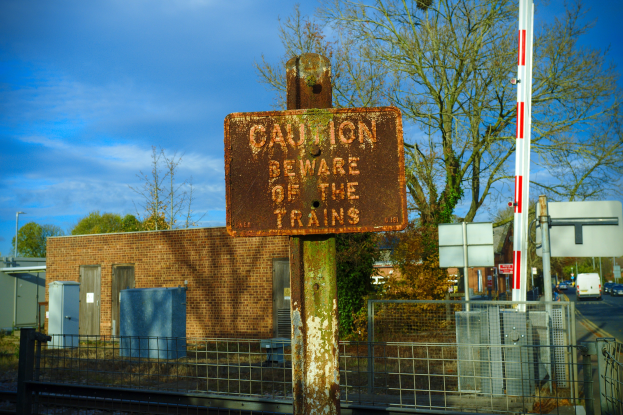 Vorsichtsschild an einem Bahnsteigzaun mit Bäumen, Pfählen, einem Gebäude, Containern, Fahrzeugen und einem bewölkten Himmel.