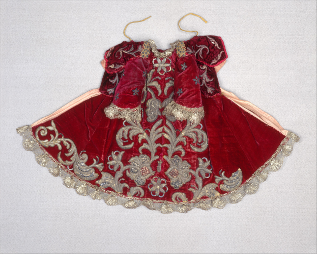 Ein rotes Samtkleid mit goldener Stickerei auf einem Bügel vor einem weißen Hintergrund, das detailreiche Verzierungen zeigt.